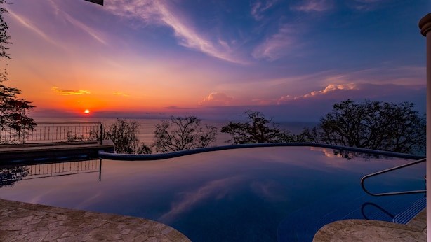 A swimming pool with a stone border and a railing, surrounded by trees and a sunset view. -  Panoramic Ocean views Waterfront Luxury Home  (Jaco)