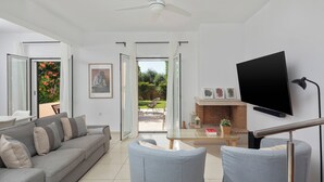 Uma sala de estar moderna com um sofá cinzento, duas poltronas, uma televisão de tela plana e um espaço de jantar com mesa e cadeiras.