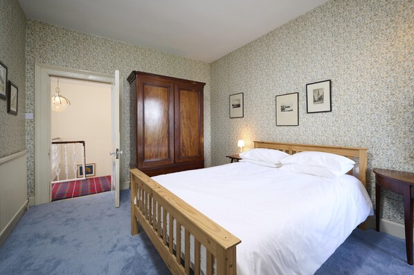 Une chambre avec un lit en bois, une table de chevet et une armoire. Une porte donne accès à une autre pièce dont le sol est recouvert d’un tapis à carreaux.