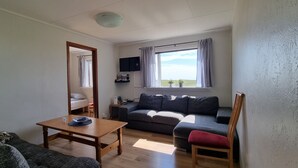 Een woonkamer met een bank, een houten tafel, een rode stoel, een televisie op een dressoir en een raam met gordijnen.