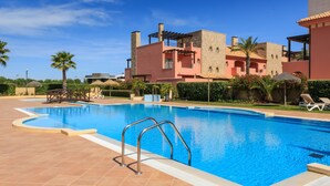 A swimming pool with metal railings, surrounded by a tiled deck. In the background, there are buildings with a mix of stone and painted exteriors, and palm trees. - Vilamoura Luxurious PENTHOUSE. 2 large pools, covered parking. (Vilamoura Portugal)