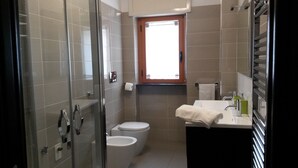 Un baño con cabina de ducha de vidrio, dos inodoros, un lavabo con espejo y una ventana que proporciona luz natural.