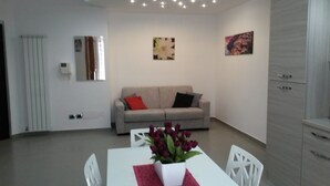 Una sala de estar moderna con un sofá gris, cojines rojo y negro, una mesa blanca con un jarrón de flores y dos cuadros colgados en la pared.