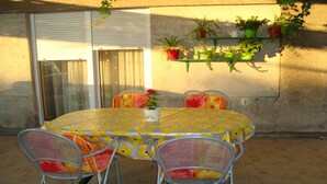 Une table ronde recouverte d’une nappe à motifs floraux jaunes, quatre chaises en métal avec des coussins rouges et une plante en pot posée sur la table.