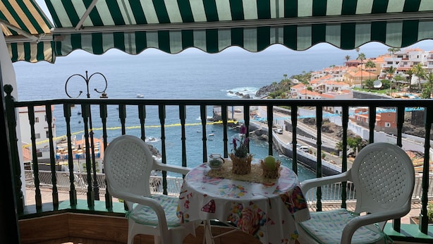 A balcony with a view of the sea and a coastal town. There are two white chairs and a small round table with a colorful tablecloth. - Playa Chica Apartment (SANTA CRUZ DE TENERIFE)