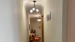 A narrow hallway with wooden flooring, a ceiling light fixture, and a door at the end leading to a room with a bed and a chair. - Playa Chica Apartment (SANTA CRUZ DE TENERIFE)