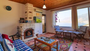 A cozy living room with a fireplace, wooden ceiling, and a large window overlooking a scenic view. - Kas Holiday Rental with Stunning Sea View and Swimming Pool ( 6 people ) (Turquoise Coast East)