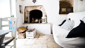 A cozy room with a fireplace, a white sofa with striped cushions, a wooden chest, and a white chair. - Authentic Trullo with swimming pool in Itria Valley Apulia (Martina Franca)