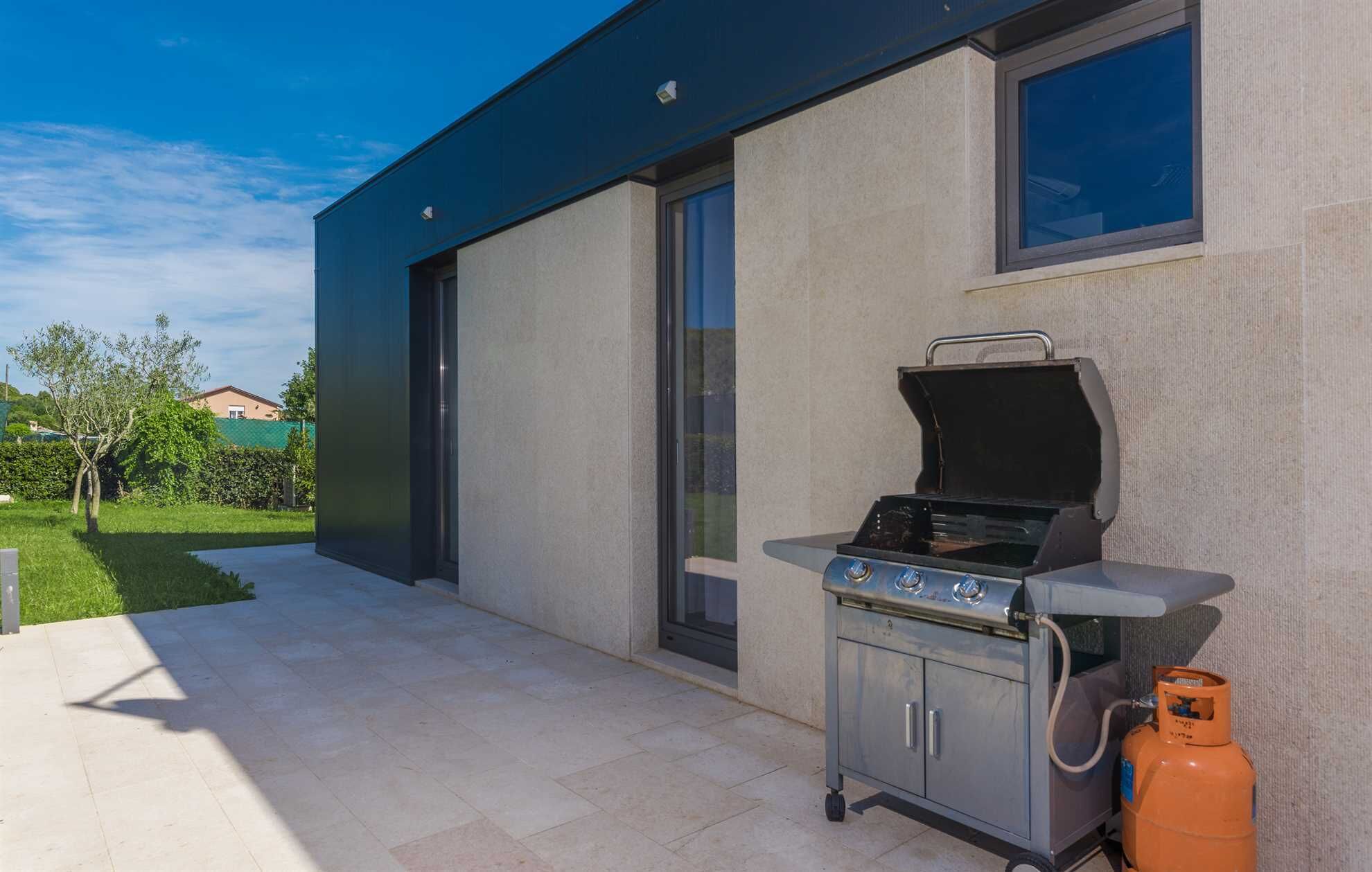 A modern outdoor kitchen with a gas grill and a propane tank, located on a patio adjacent to a building with large windows.