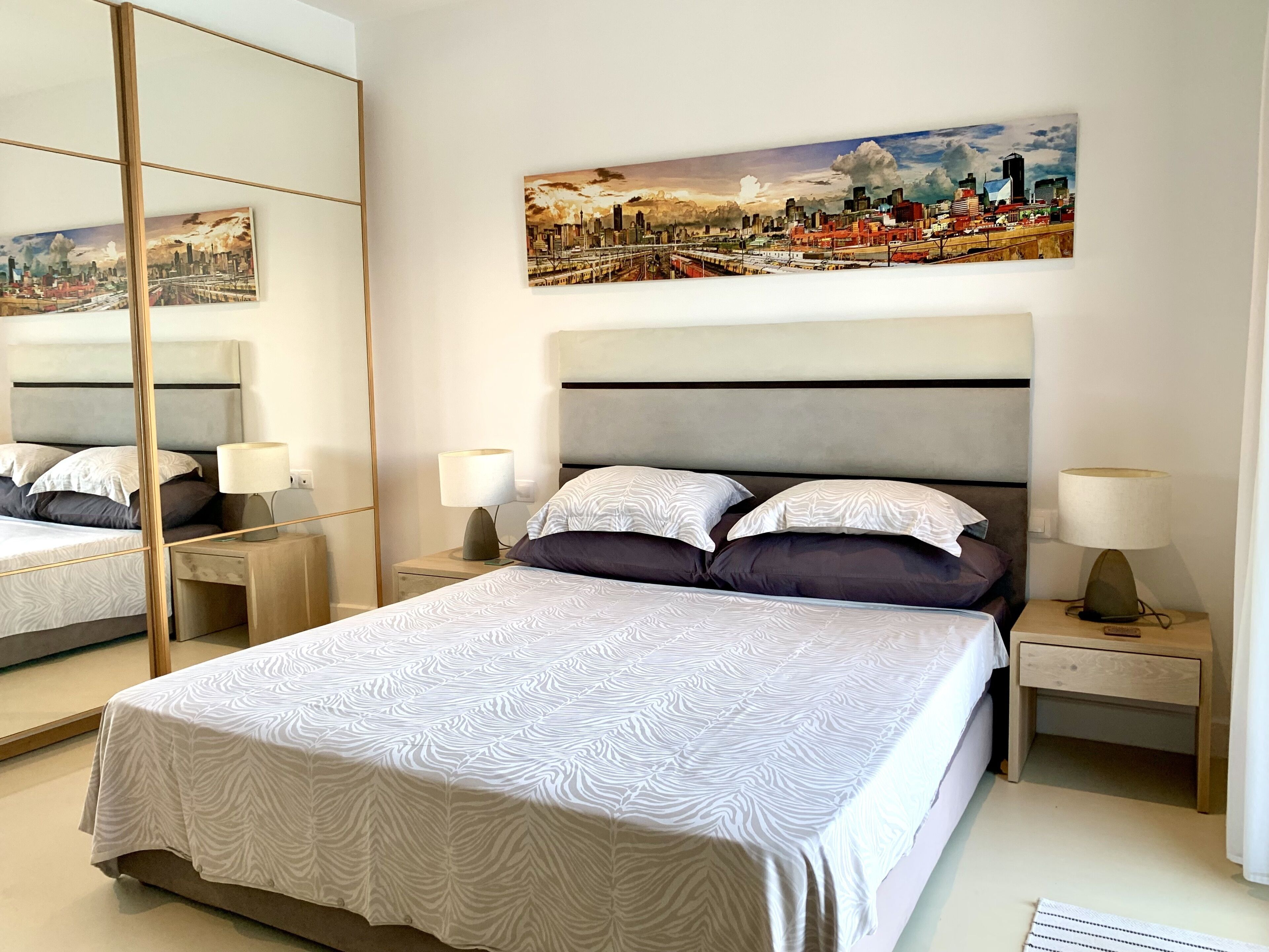 A modern hotel room with a large bed, two bedside tables, a mirror, and a cityscape painting on the wall.