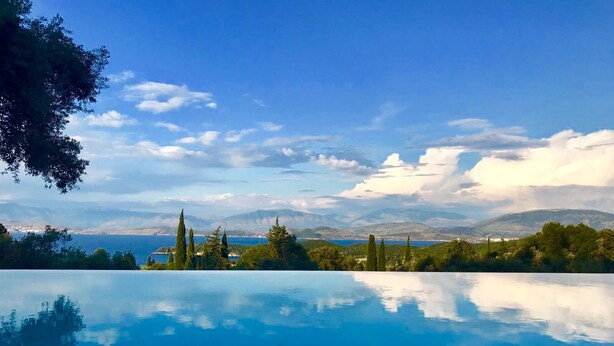 A clear blue sky, a calm pool reflecting the sky and trees, and a distant view of mountains and a body of water.