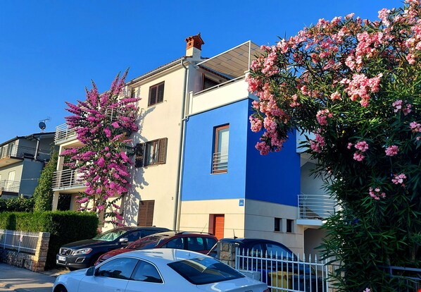A residential building with balconies, parked cars, and flowering trees.