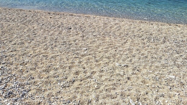 A pebble beach with clear blue water and a distant sailboat.