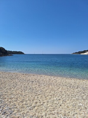 A pebble beach with clear blue water and a distant sailboat.