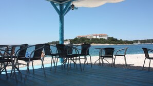 A beachside patio with metal chairs and tables, overlooking a calm sea and a building with a red roof.