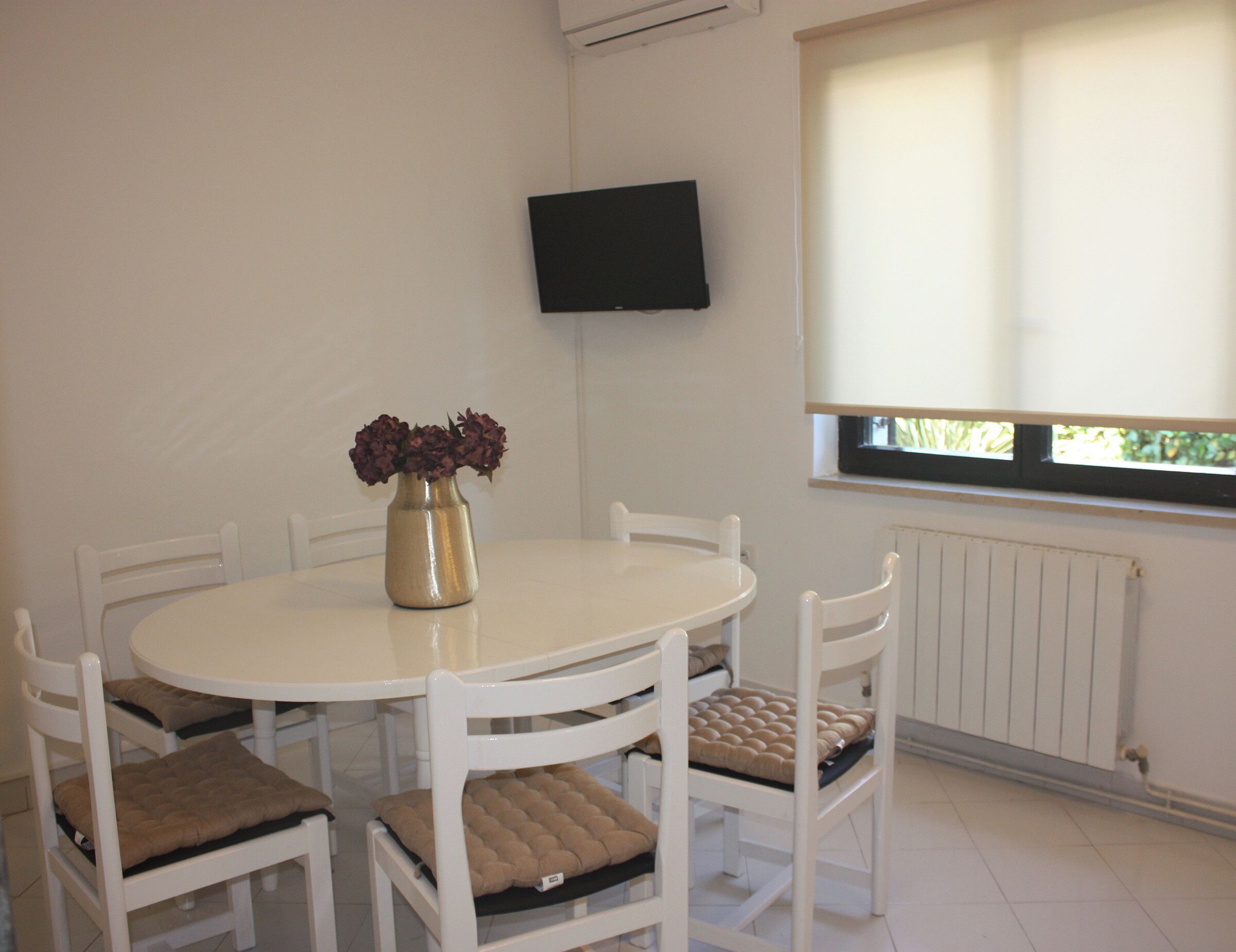 Un coin repas avec une table ronde, des chaises blanches, un vase de fleurs, un téléviseur fixé au mur et une fenêtre avec des stores.