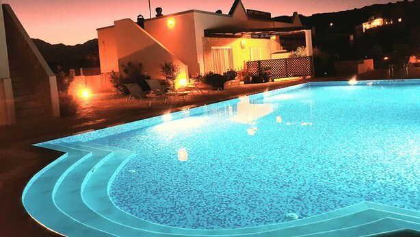 Ein Pool mit geometrischem Design, umgeben von einer Terrasse mit Liegestühlen und einem Gebäude mit geneigtem Dach im Hintergrund.