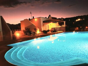 Una piscina con diseño geométrico, rodeada de un patio con sillones de descanso y un edificio con techo inclinado al fondo.
