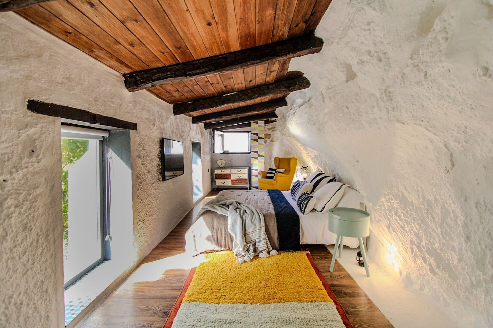 Un dormitorio rústico y estrecho con techo de madera, una cama con alfombra a rayas amarillas y blancas, un sillón amarillo y una mesita de noche con una lámpara.