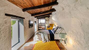 Un dormitorio rústico y estrecho con techo de madera, una cama con alfombra a rayas amarillas y blancas, un sillón amarillo y una mesita de noche con una lámpara.