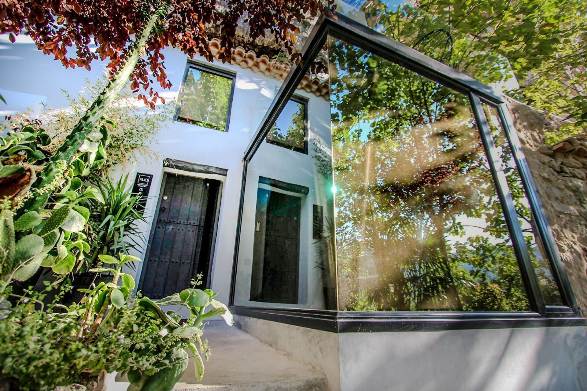Una casa moderna con fachada de vidrio, una puerta y un pequeño jardín con diversas plantas.