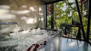 Un espace spa agrémenté d’un élément aquatique et de pétales de rose, de grandes fenêtres offrant une vue sur les arbres et un mur en béton.