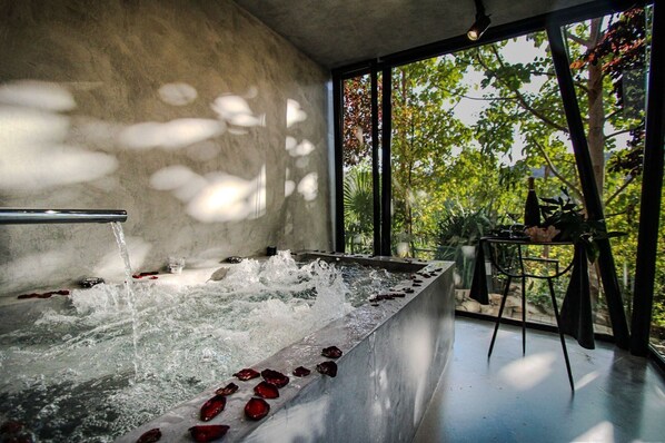Un espace spa agrémenté d’un élément aquatique et de pétales de rose, de grandes fenêtres offrant une vue sur les arbres et un mur en béton.
