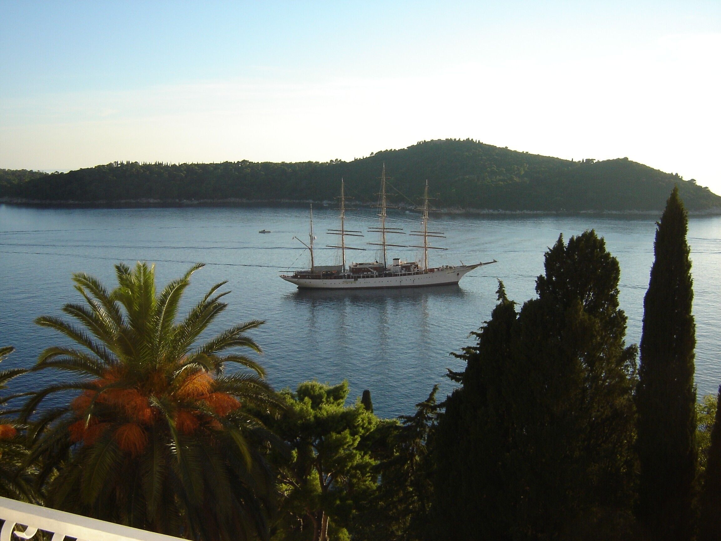 A large sailing ship on calm waters with a forested hill in the background.