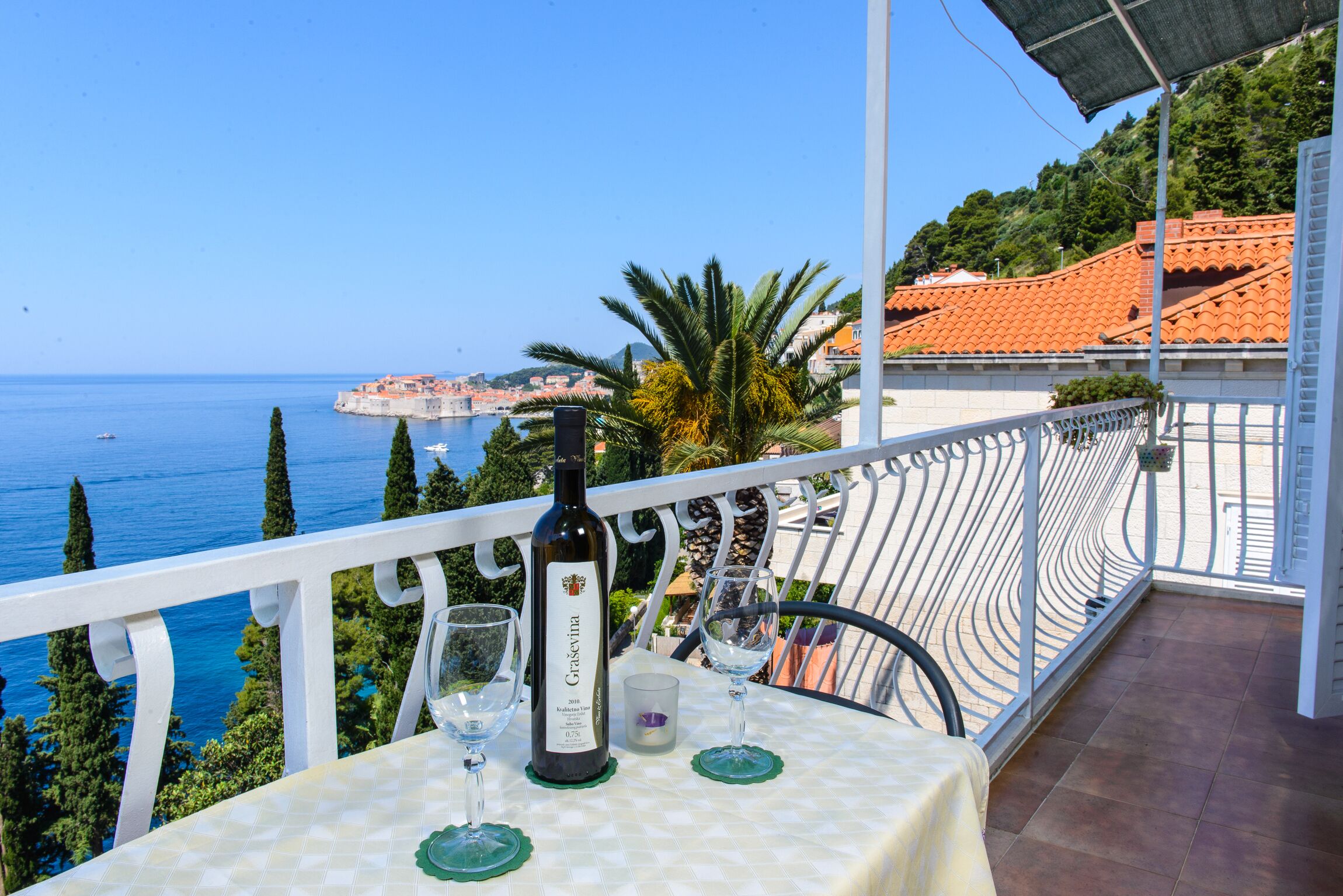 Een tafel met een fles wijn, twee glazen en een kaars op een balkon met uitzicht op zee en een kustplaats.