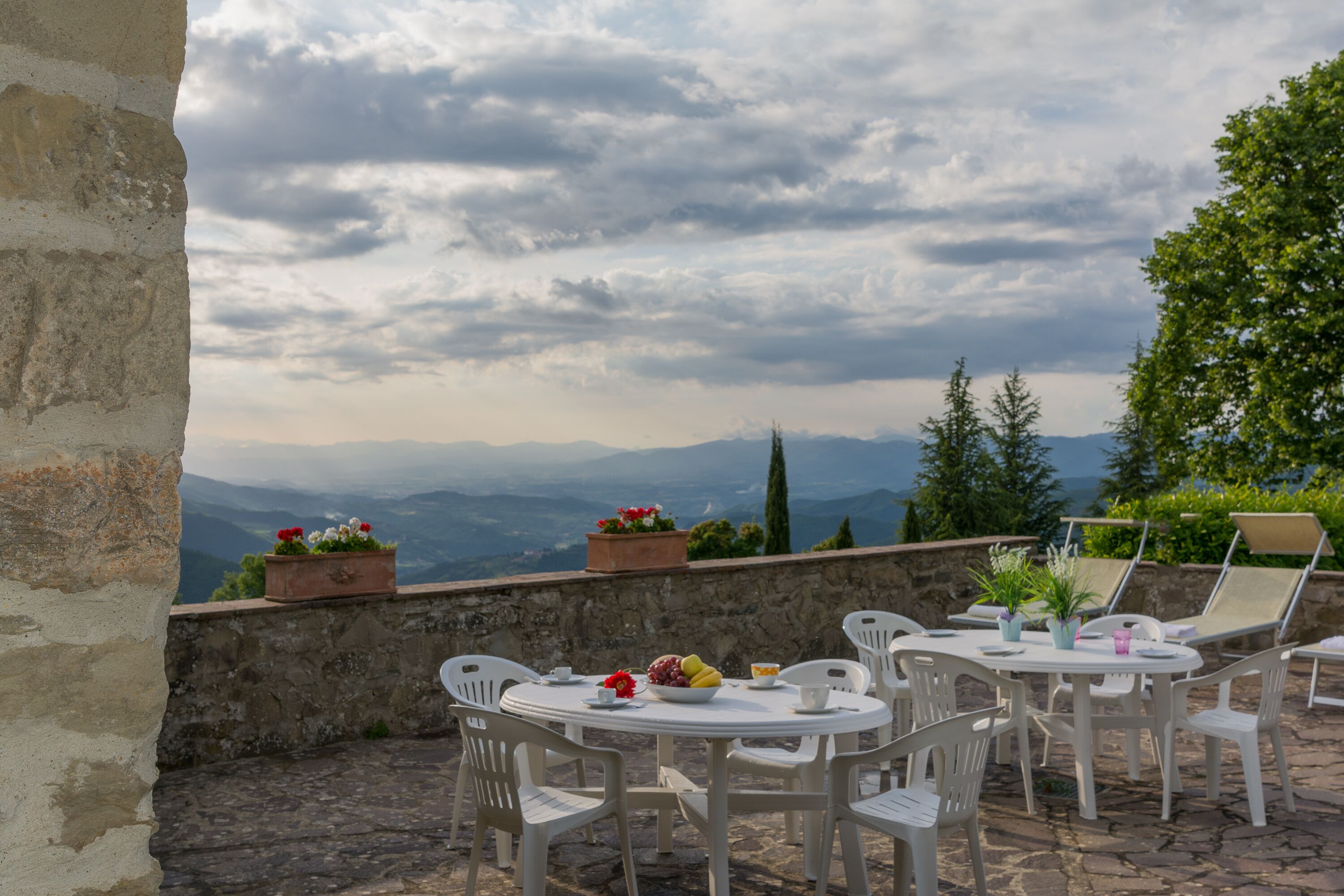 Eine Terrasse im Freien mit einer Steinmauer, weißen Stühlen, einem runden Tisch und Blumentöpfen.