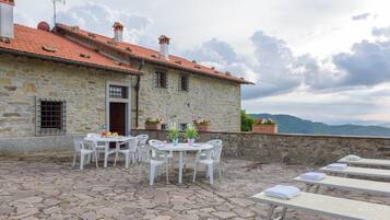 Un edificio in pietra con tetto in tegole, area esterna con sedie e tavoli bianchi e fioriere sulla parete esterna.