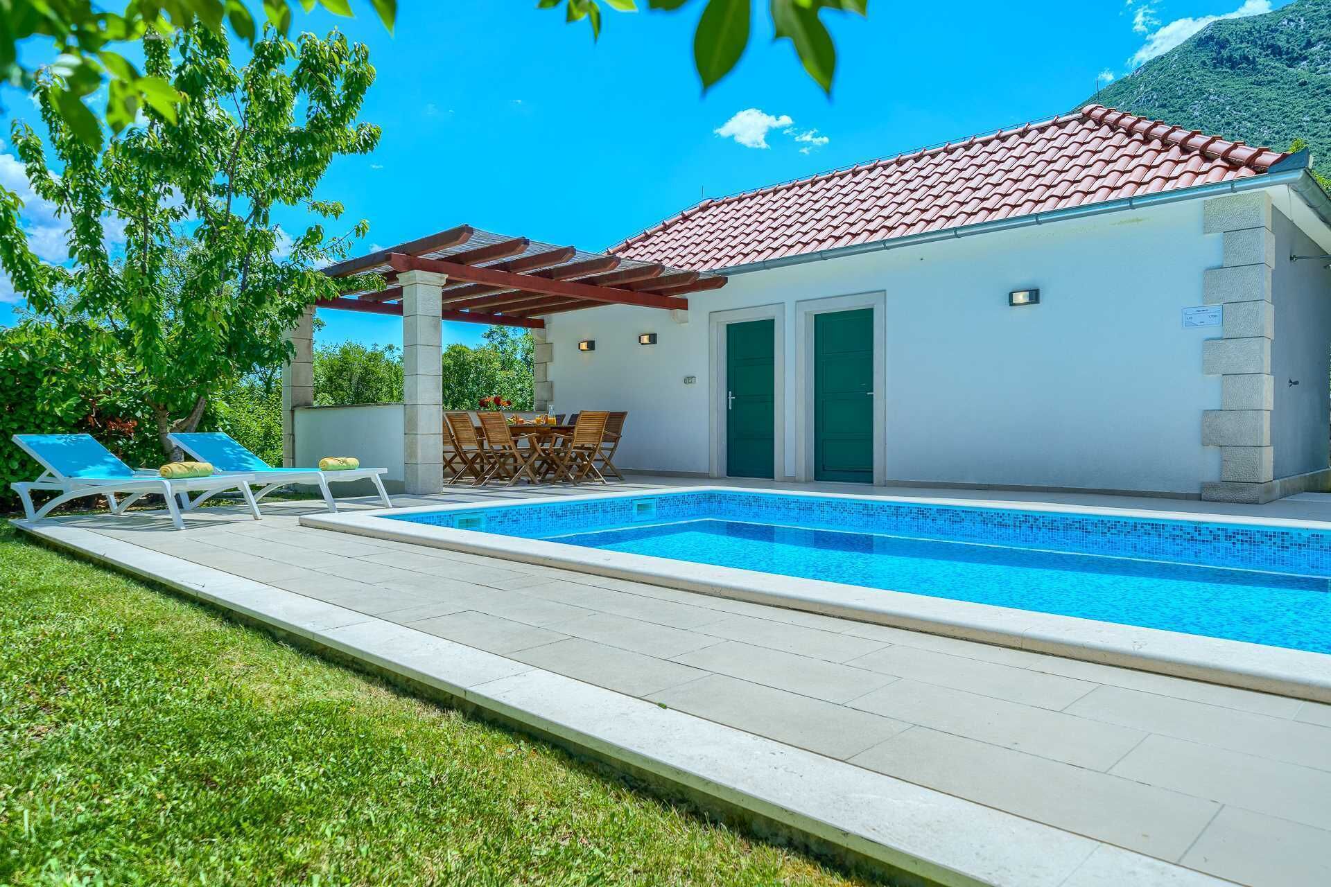 Un área junto a la piscina con un suelo de baldosas, dos sillones de descanso, una mesa con sillas y una pérgola.