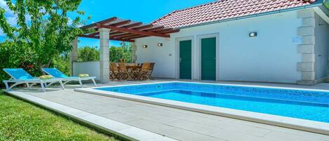 Un área junto a la piscina con un suelo de baldosas, dos sillones de descanso, una mesa con sillas y una pérgola.