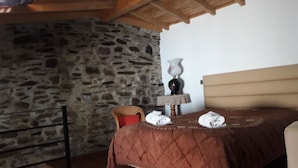 A bedroom with a stone wall, a wooden ceiling, a bed with a brown quilt, a small table with a lamp, and a chair.