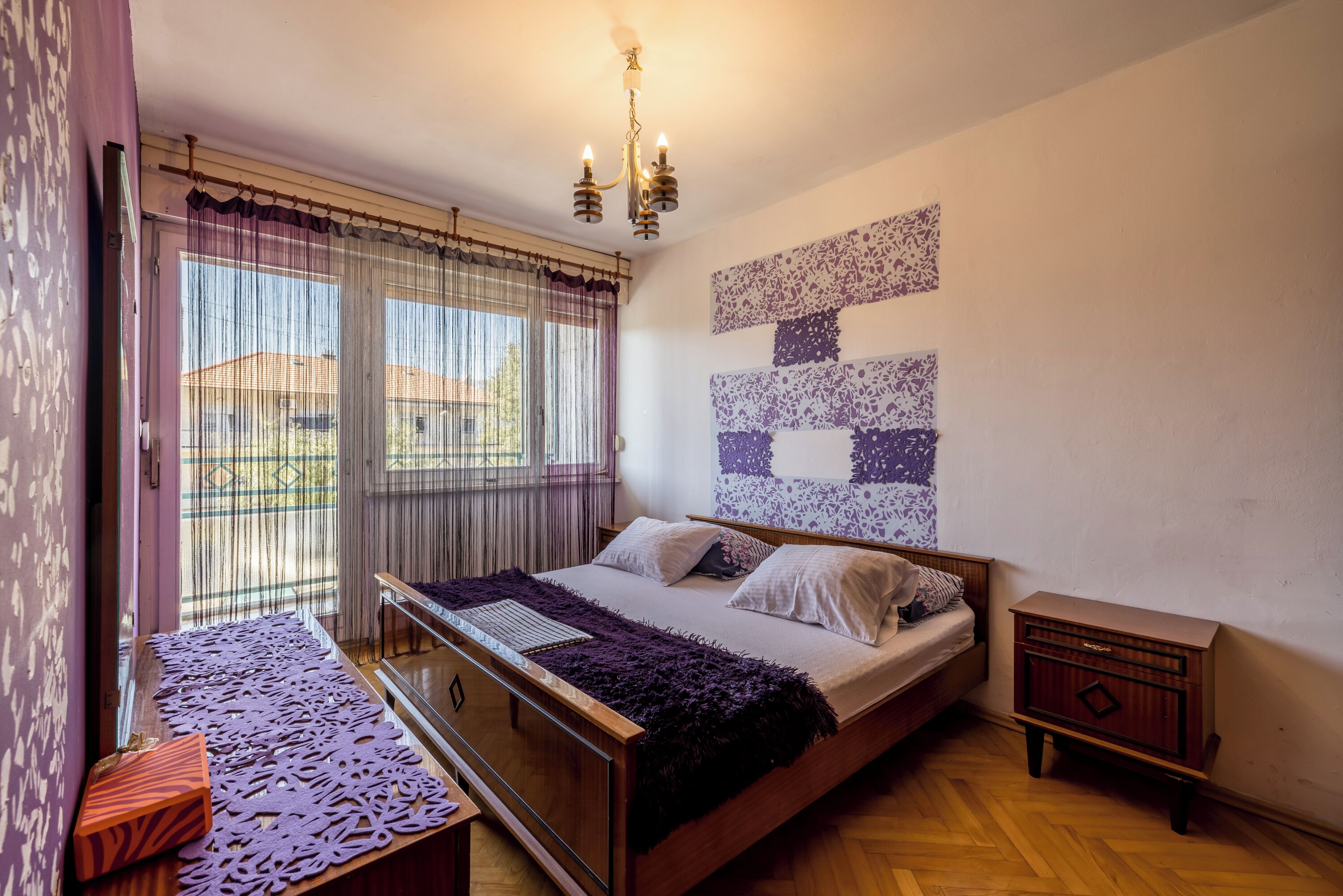 A bedroom with a bed, a bedside table, a nightstand, a window with curtains, and a wall with a patterned design.
