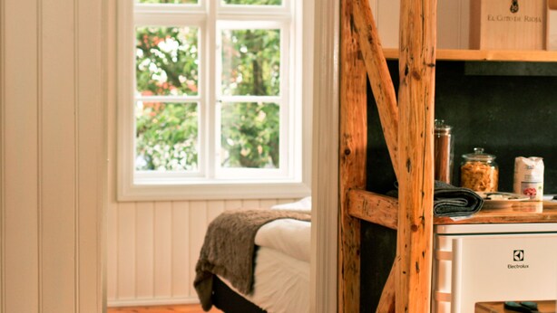 Un dormitorio con estructura de cama de madera, una ventana con vista a follaje y una zona de cocina con mostrador y una bolsa con el texto 'El Gato de Rojo'.