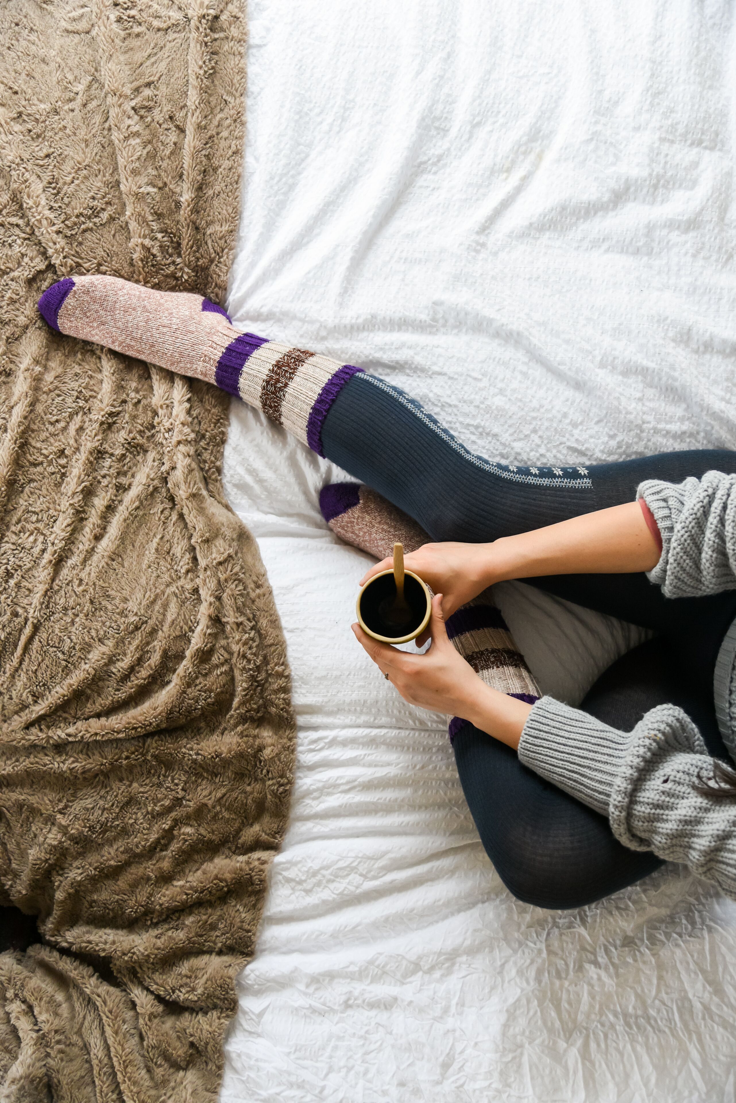 Una persona sentada en una cama con una manta marrón, sosteniendo una taza.