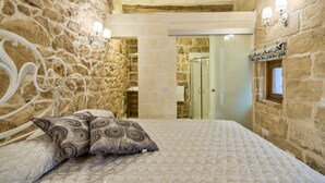 A bedroom with stone walls, a bed with patterned pillows, a wooden headboard, and a glass shower door. - Highly Renovated Ta Rozamari Character House With Pool And Hot Tub (Zejtun)