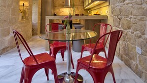 A stone-walled dining area with a glass table and red chairs, a kitchen in the background, and wooden beams on the ceiling. - Highly Renovated Ta Rozamari Character House With Pool And Hot Tub (Zejtun)