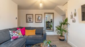 Ein Wohnzimmer mit grauem Sofa, bunten Kissen, einer hängenden Leuchte, gerahmten Bildern, einem Spiegel und Topfpflanzen.