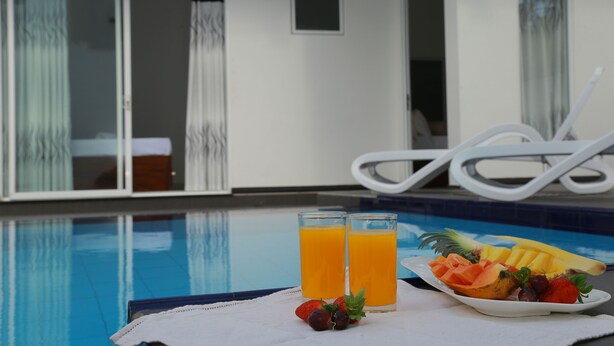 Escena a la piscina amb una taula que té dos vasos de suc de taronja, un plat de fruita i una tovallola.
