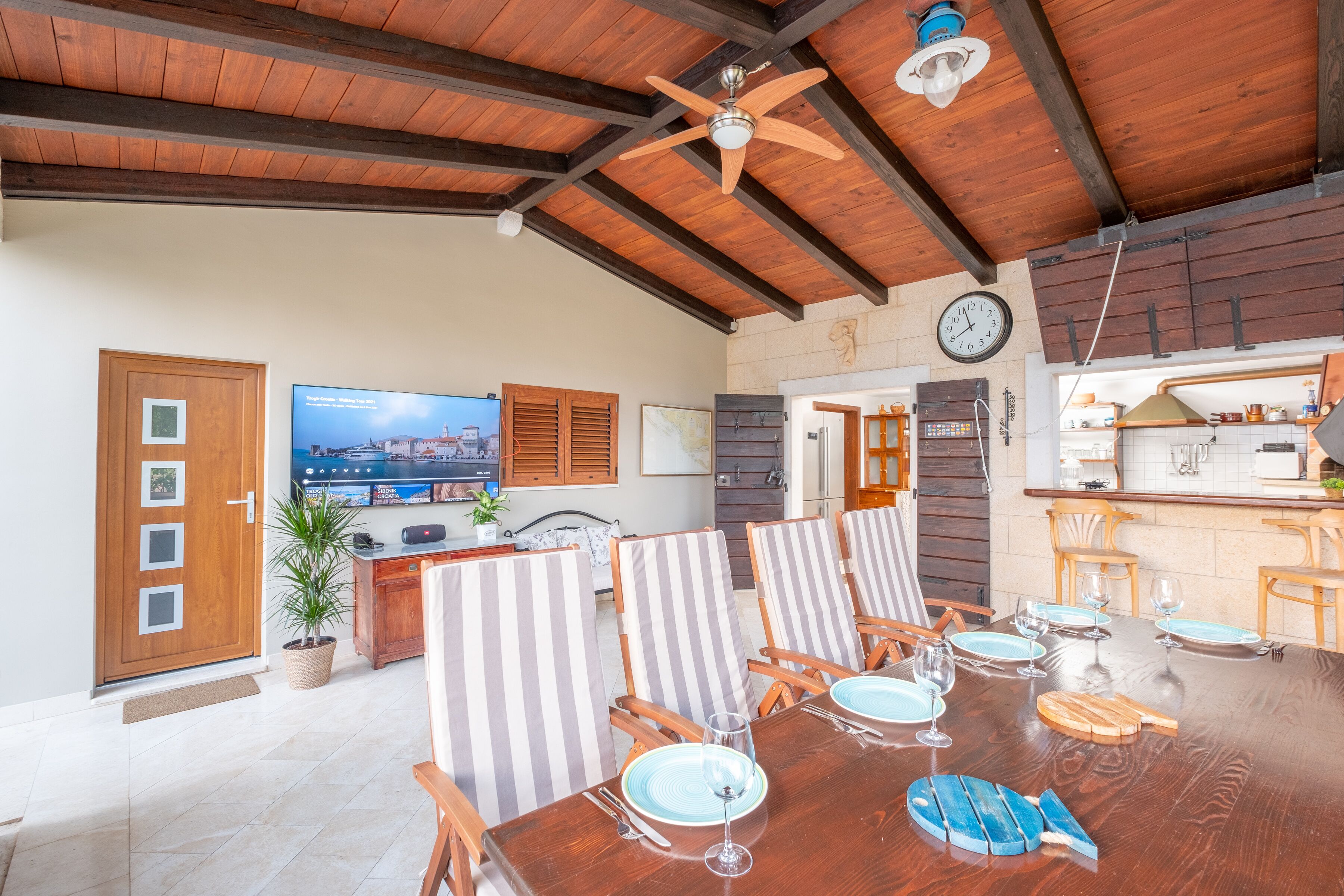 Un coin repas avec une table en bois dressée pour six personnes, des chaises à rayures, un ventilateur de plafond et une cuisine en arrière-plan.