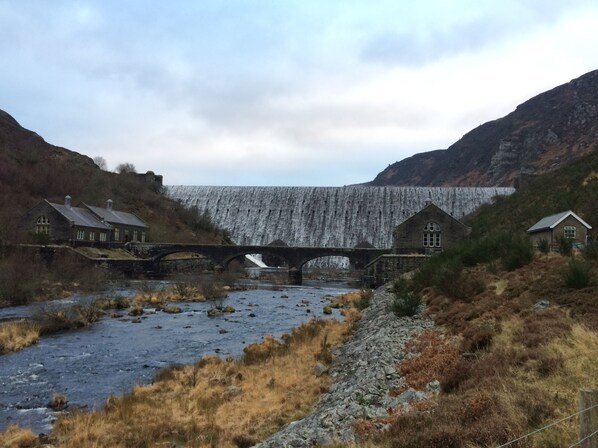 A stone bridge over a river with houses on either side and a dam in the background. - Gwardolau Cottage, Wye Valley Retreat. (Rhayader)