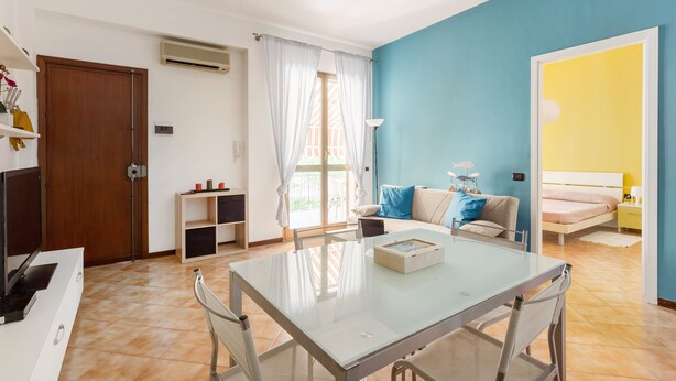 Un salon moderne avec une table à manger en verre, un canapé avec des coussins bleus et un lit avec une tête de lit jaune.