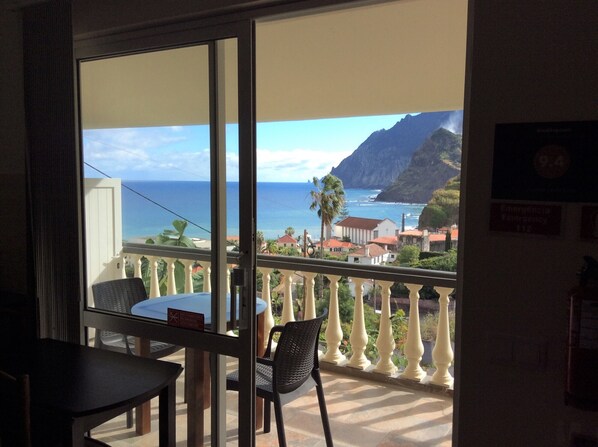 A balcony with a table and chairs, overlooking a coastal view with buildings and mountains.
