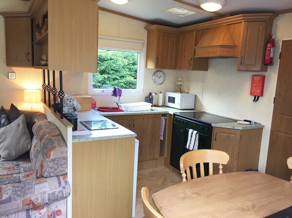 A compact kitchenette with wooden cabinets, a microwave, a stove, and a sink. There is a window providing natural light, and a fire extinguisher mounted on the wall. - Enjoy a break in our dog friendly, chalet style, static van with ferry discounts (St. Helens, Isle of Wight)