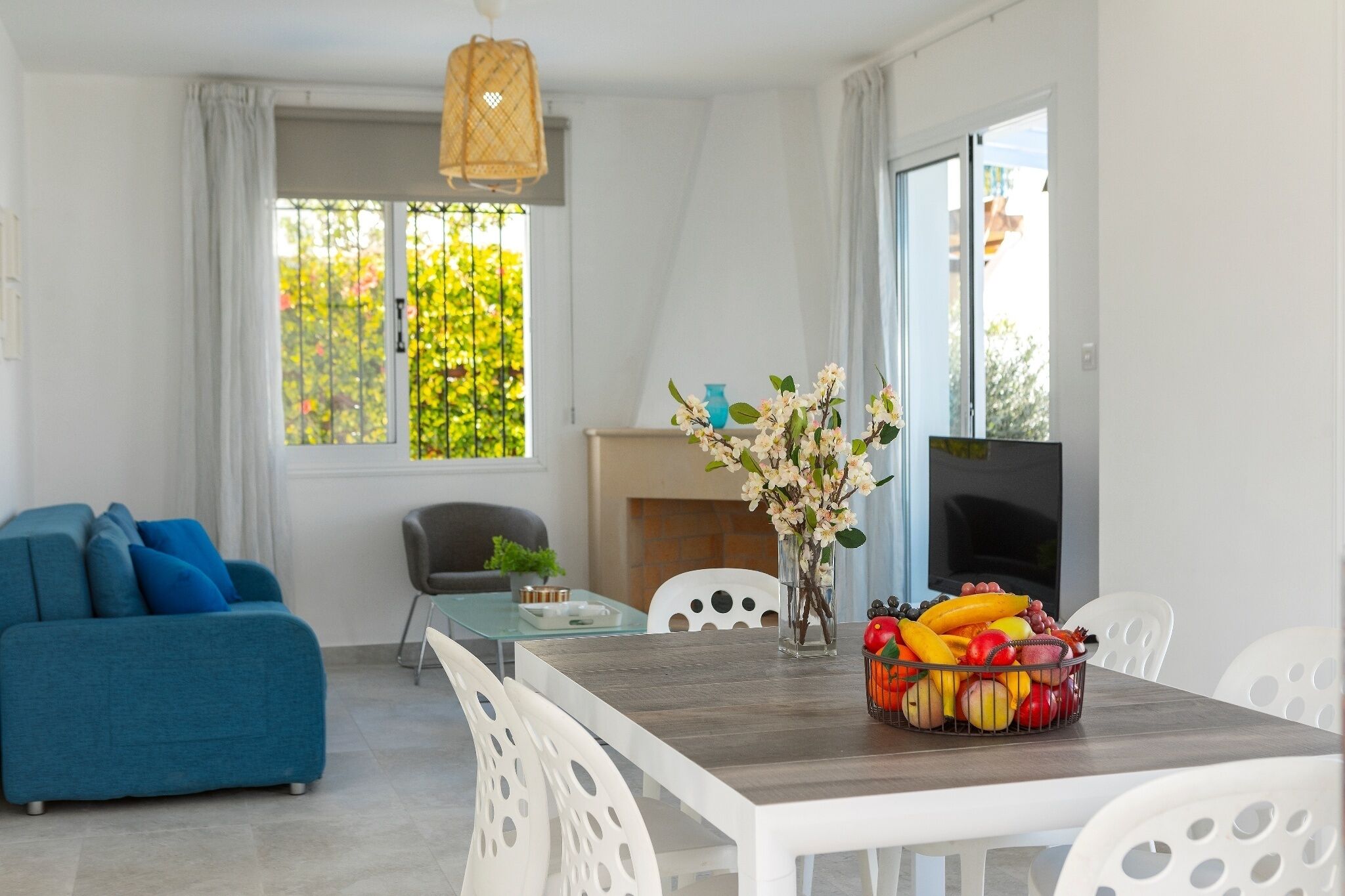 A modern living room with a sofa, dining table, and chairs. There is a TV, a fireplace, and a window with a view of greenery.