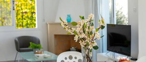 A modern living room with a glass coffee table, a vase with flowers, a fruit bowl, and a television.