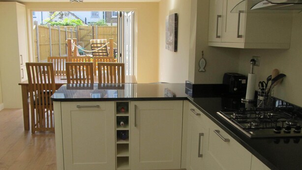 A kitchen with wooden cabinets, a black countertop, and a gas stove. There is a dining area with wooden chairs and a table visible through an open door. - Chloe Cottage - Family friendly fishermans cottage close to the beach and harbou (Whitstable)