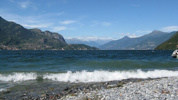 Vista costeira com praia rochosa, mar calmo e montanhas ao fundo.